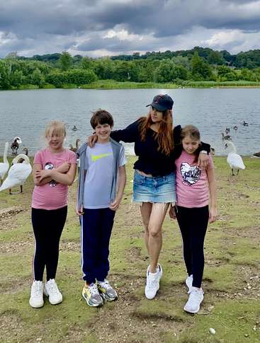 Das Bild zeigt eine Frau und zwei Kinder, die im Gras vor einem See stehen, mit Schwänen und Enten im Hintergrund, unter einem bewölkten Himmel.