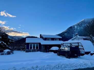 Uma casa fica cercada por neve profunda, com um caminhão e um trailer na garagem. Ao fundo, dá pra ver montanhas cobertas de neve e um céu azul claro.