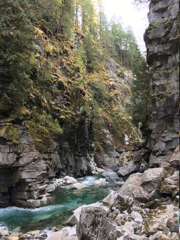 Um riacho azul e cristalino serpenteia por um desfiladeiro rochoso com penhascos íngremes e escarpados, cobertos de musgo e árvores perenes, criando uma paisagem natural e tranquila.