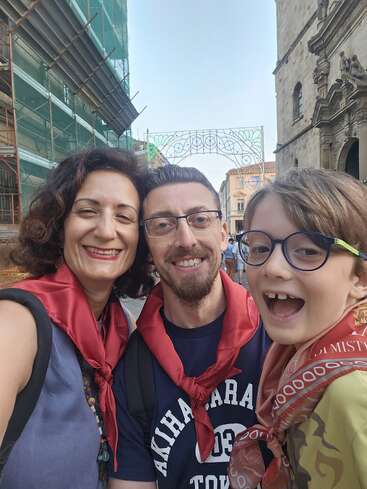 A smiling family of three poses closely together on a street, all wearing red scarves. They appear happy, enjoying a festive day in an urban setting.
