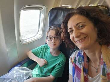 A woman and a young boy, both smiling, sit together on an airplane by the window, ready for a journey. The boy wears glasses and a green shirt.