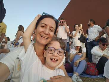 A joyful woman and child take a happy selfie at an outdoor event, surrounded by a crowd of people, all casually dressed, enjoying the day.