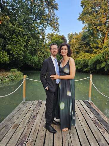 A couple dressed elegantly, stands on a wooden dock over a calm green pond, surrounded by lush trees, smiling warmly on a bright, sunny day.