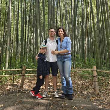 A family of three stands together in a scenic bamboo forest. They smile, posing for a photo on a path surrounded by tall, green bamboo stalks.