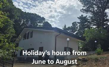 A white holiday house surrounded by trees under a partly cloudy sky. Outdoor seating, lush green garden, and a serene environment, available from June to August.