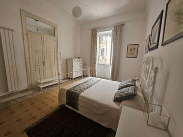 Ein helles, gemütliches Schlafzimmer mit einem Doppelbett, gemusterter Bettwäsche, einem eleganten Eisengestell, einer Kommode, einem Fenster mit Vorhängen, Wandkunst und einem klassischen Fliesenboden.