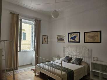 Ein ruhiges Schlafzimmer mit einem weißen Metallbett, heller Bettwäsche mit gemusterten Akzenten, zwei Nachttischen, einem Fenster mit beigen Vorhängen und Kunstwerken an den weißen Wänden.