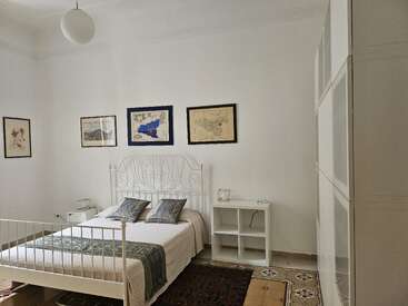 Ein helles, minimalistisches Schlafzimmer mit einem weißen, verzierten Bett, zwei Nachttischen, gerahmten Landkarten an der Wand, gefliestem Boden, gemustertem Teppich und einem großen Kleiderschrank.