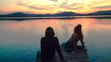 Two people sit on a wooden dock, enjoying a tranquil lake at sunset. The sky glows with warm colors reflecting peacefully on the calm water.