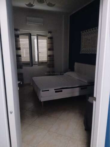 Um quarto arrumado com cama de casal, cortinas listradas, janela grande, ar condicionado, piso de azulejo, parede azul escura e uma decoração na parede.