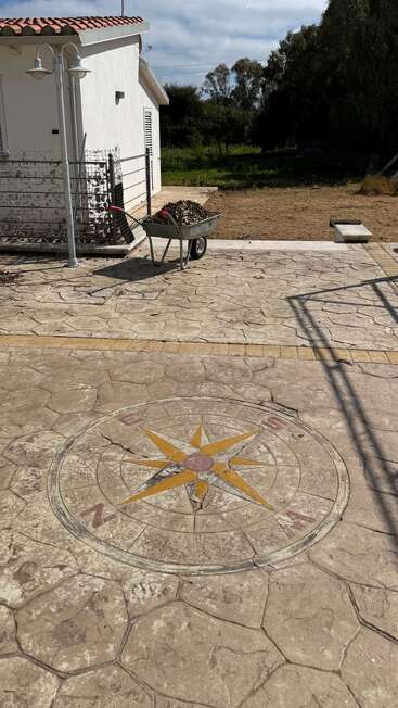 Ein weißes Haus mit Ziegeldach steht neben einem Garten. Man sieht eine Schubkarre mit Laub, einen Gehweg mit einem Kompassrosenmuster und einen Laternenpfahl.