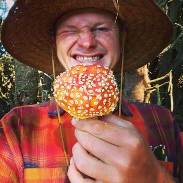 La imagen muestra a un hombre con sombrero de paja y camisa de cuadros, sosteniendo una seta con manchas blancas sobre un gorro naranja, sobre un fondo boscoso.