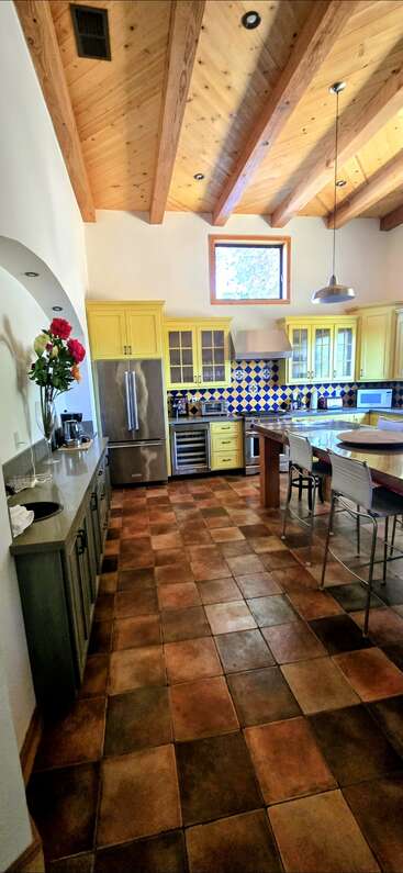 Esta cocina cuenta con armarios amarillos, techo con vigas de madera, salpicadero de azulejos de colores, baldosas de terracota, electrodomésticos de acero inoxidable, isla con taburetes y un jarrón de flores.