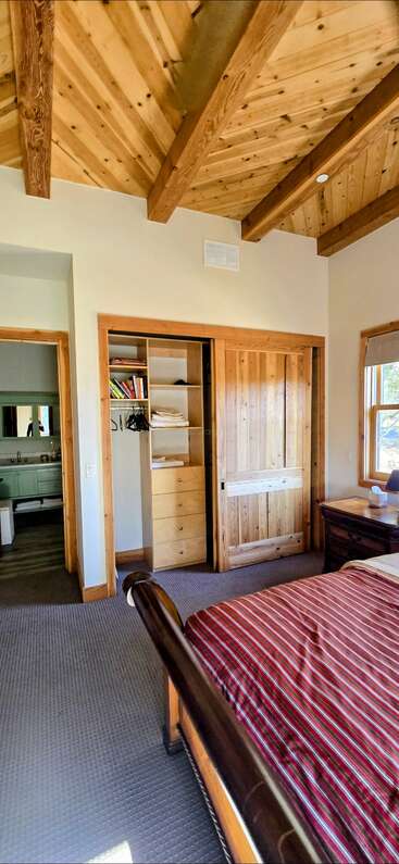 Este acogedor dormitorio presenta un techo de madera con vigas vistas, un armario abierto, una colcha de rayas, suelo enmoquetado y un cuarto de baño adyacente visible a través de la puerta.