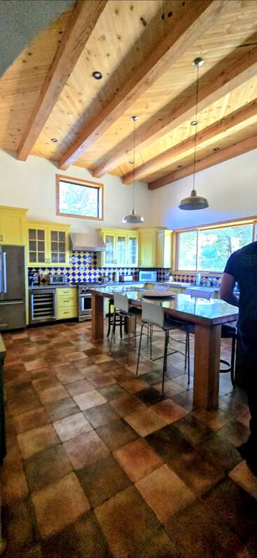 Esta cocina cuenta con armarios amarillos, salpicadero de azulejos azules y amarillos, techo con vigas de madera, isla grande, lámparas colgantes, suelo de baldosas, electrodomésticos inoxidables y mucha luz natural.