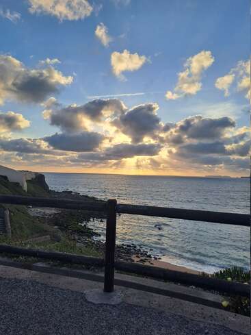 La imagen capta una impresionante puesta de sol junto al mar, con rayos de sol que se abren paso entre nubes dispersas, iluminando el océano. En primer plano aparecen una valla y una costa rocosa.