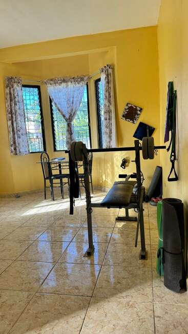 In einem sonnenbeschienenen Raum mit gelben Wänden stehen eine Hantelbank, Trainingsgeräte, Matten und ein kleiner Esstisch an drei großen, mit Vorhängen versehenen Fenstern. Gemütliche Atmosphäre.