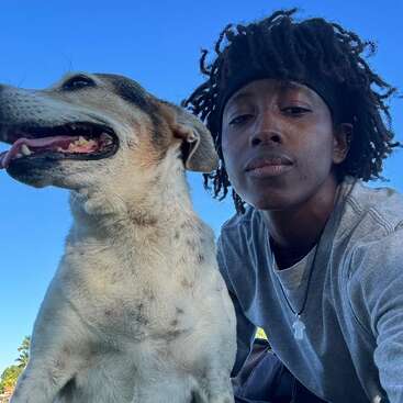 Eine Person mit Dreadlocks, die ein graues Hemd und eine Halskette trägt, posiert neben einem glücklichen Hund im Freien. Der Himmel ist strahlend blau und schafft eine fröhliche Atmosphäre.