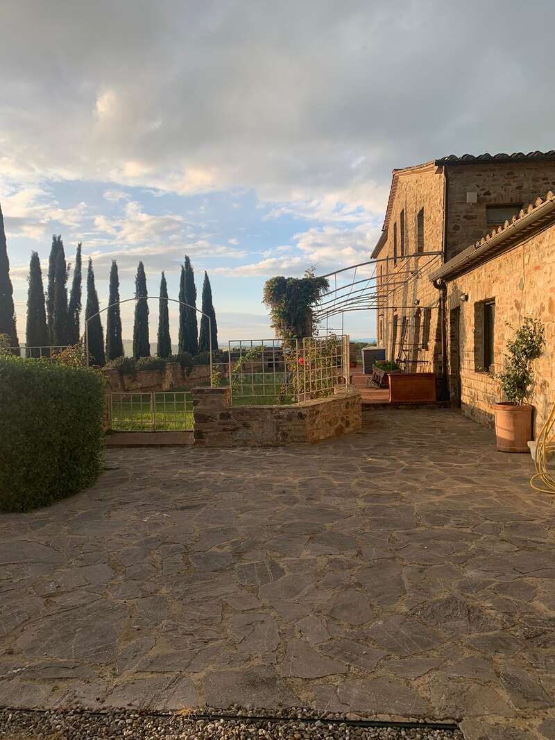 A imagem retrata um edifício de pedra sereno com um jardim, com um caminho de pedra, treliça em arco e um céu nublado, evocando um ambiente rural tranquilo.