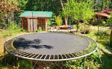 A large round trampoline sits outdoors in a green garden, surrounded by trees and wire fencing. In the background, there are two wooden buildings.