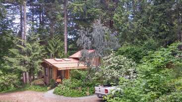 A cozy wooden cabin is nestled among lush green trees and dense foliage. A white pickup truck is parked on a gravel driveway in the peaceful forest.
