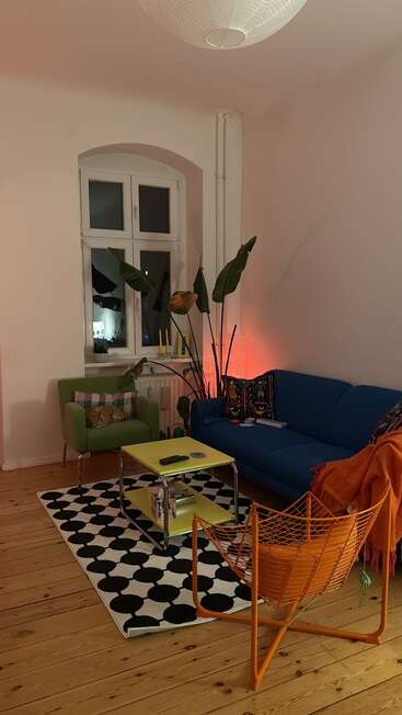A cozy living room featuring a blue sofa, green armchair, orange chair, patterned rug, yellow table, large plant, and ambient lighting, with wooden floors.