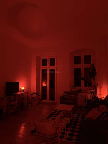 A cozy living room is bathed in red light, featuring large windows, a sofa, plants, television, modern furniture, and a patterned rug on wooden floors.