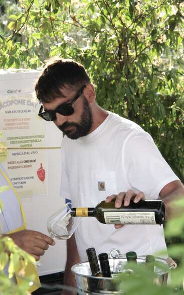 Um homem usando óculos escuros e camiseta branca serve vinho branco em uma taça ao ar livre, cercado por vegetação, com um pôster visível ao fundo.