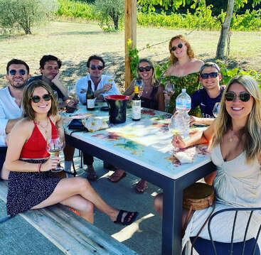 A group of friends sit around a colorful outdoor table, enjoying wine and smiles on a sunny day, surrounded by green vines and nature.