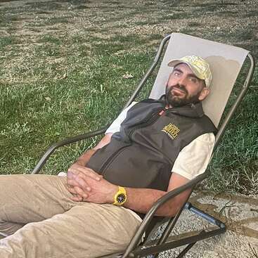 Un hombre con barba se relaja en una tumbona al aire libre, con gorra, chaleco oscuro, pantalones beige y reloj amarillo, disfrutando de su entorno.