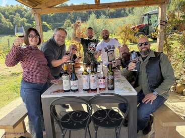 Eine Gruppe von Freunden genießt gemeinsam Wein unter einer hölzernen Pergola, lächelt und stößt mit Gläsern an, umgeben von Weinbergen und malerischer Natur.