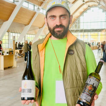 A bearded man in a green shirt and cap stands indoors, holding two wine bottles. The background features people, wooden beams, and large windows with sunlight.
