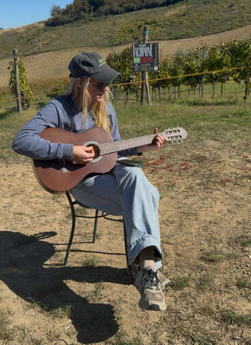 A person wearing sunglasses and a cap sits on a chair outside, playing a guitar. Behind them are green vineyards, a sign, and sunlit hills.