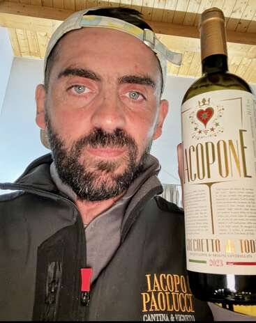 A bearded man wearing a cap and a branded jacket holds a wine bottle labeled "Iacopone." He stands indoors under a wooden ceiling, looking directly at the camera.
