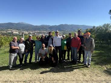 A group of people pose outdoors in a vineyard with mountains in the background, under a clear blue sky, enjoying a sunny day together, smiling.