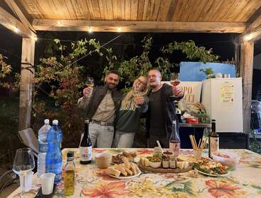 Three people smile and toast with wine glasses under a wooden gazebo at night. The table is set with bread, appetizers, drinks, and festive lights.