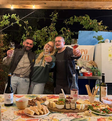 Three friends stand together, smiling and holding glasses of wine, enjoying a festive outdoor dinner. The table is filled with bread, cheese, salads, and wine bottles.