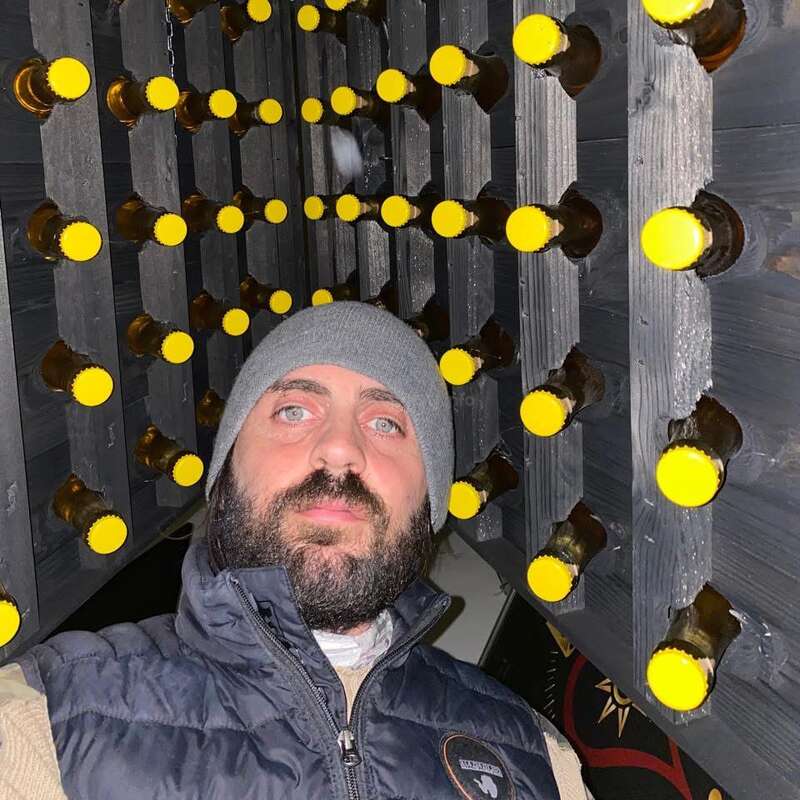 A bearded man in a gray beanie and vest stands beneath a rack of beer bottles with yellow caps, creatively displayed horizontally on black wooden shelves.