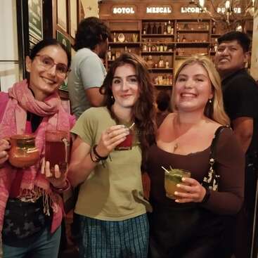 Three smiling women holding colorful cocktails pose together in a cozy bar with wooden shelves and bottles behind them. Other patrons and warm lighting complete the lively atmosphere.