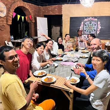 The image depicts a diverse group of people gathered around a long table, enjoying a meal together in a warm and inviting setting.