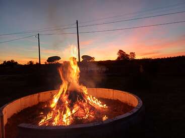 Una gran hoguera arde dentro de un pozo circular al anochecer, con llamas vibrantes que iluminan el paisaje que se oscurece y el colorido cielo del atardecer al fondo.
