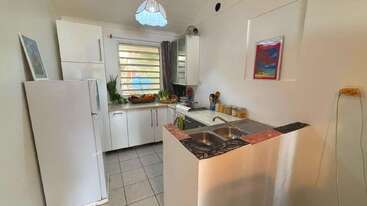 This cozy kitchen features white cabinets, a refrigerator, a double sink, tiled floor, window with blinds, countertop fruit, wall art, and a ceiling light fixture.
