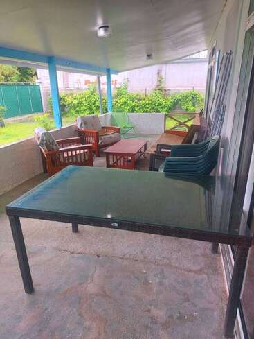 This image shows a covered outdoor patio with cushioned chairs, sofas, a glass-top table, plastic chairs, and greenery in the yard. The atmosphere is relaxed.