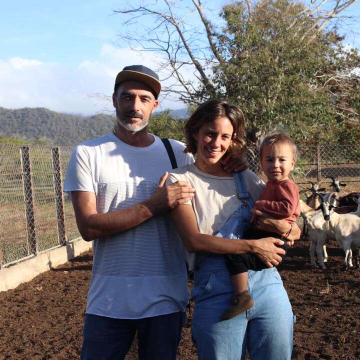 The image depicts a family of three posing for a photo in front of a fence, with two white goats standing behind them in a dirt enclosure on a sunny day.