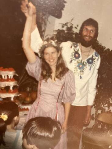 A happy couple celebrates at a party or wedding, lifting their hands in joy. The woman wears a floral crown, surrounded by cake and children.