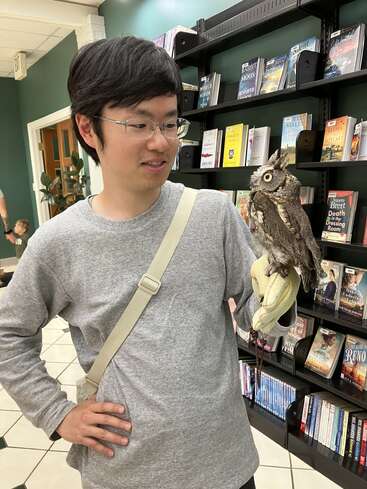 A man wearing glasses and a gray shirt holds a small owl on his gloved hand, standing in a bookshop with shelves full of books.