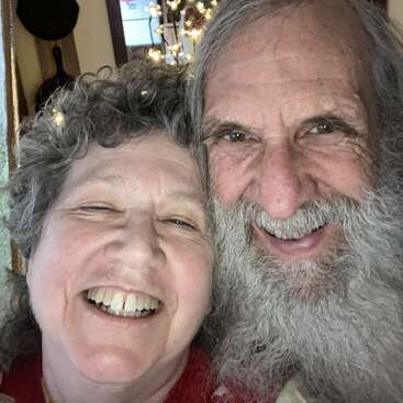 A joyful elderly couple smiles warmly in a close-up selfie, their faces radiating happiness and affection, surrounded by a cozy indoor background with soft lighting.