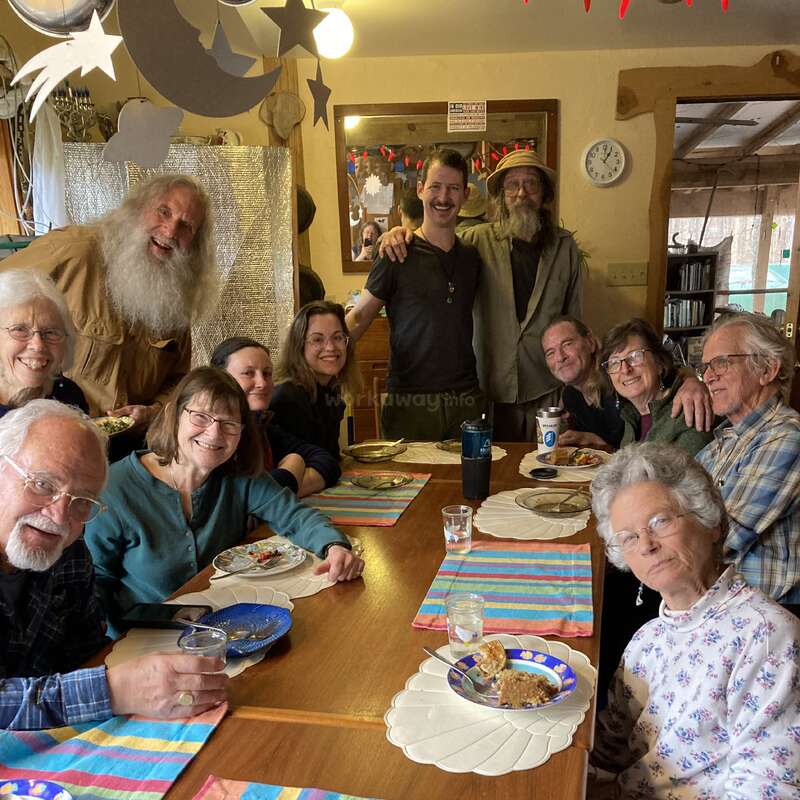 A group of twelve smiling people sit and stand around a wooden table with colorful placemats, sharing a meal in a cozy, decorated room.