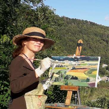 Una mujer con un sombrero de paja pinta un colorido paisaje al aire libre. Sonríe, con guantes y delantal, rodeada de vegetación y colinas en la lejanía.