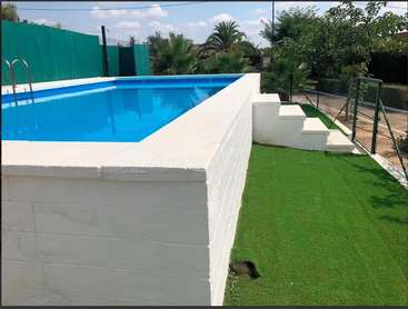 The image depicts a rectangular swimming pool with a white wall and steps, surrounded by artificial grass and trees, set against a blue sky with clouds.